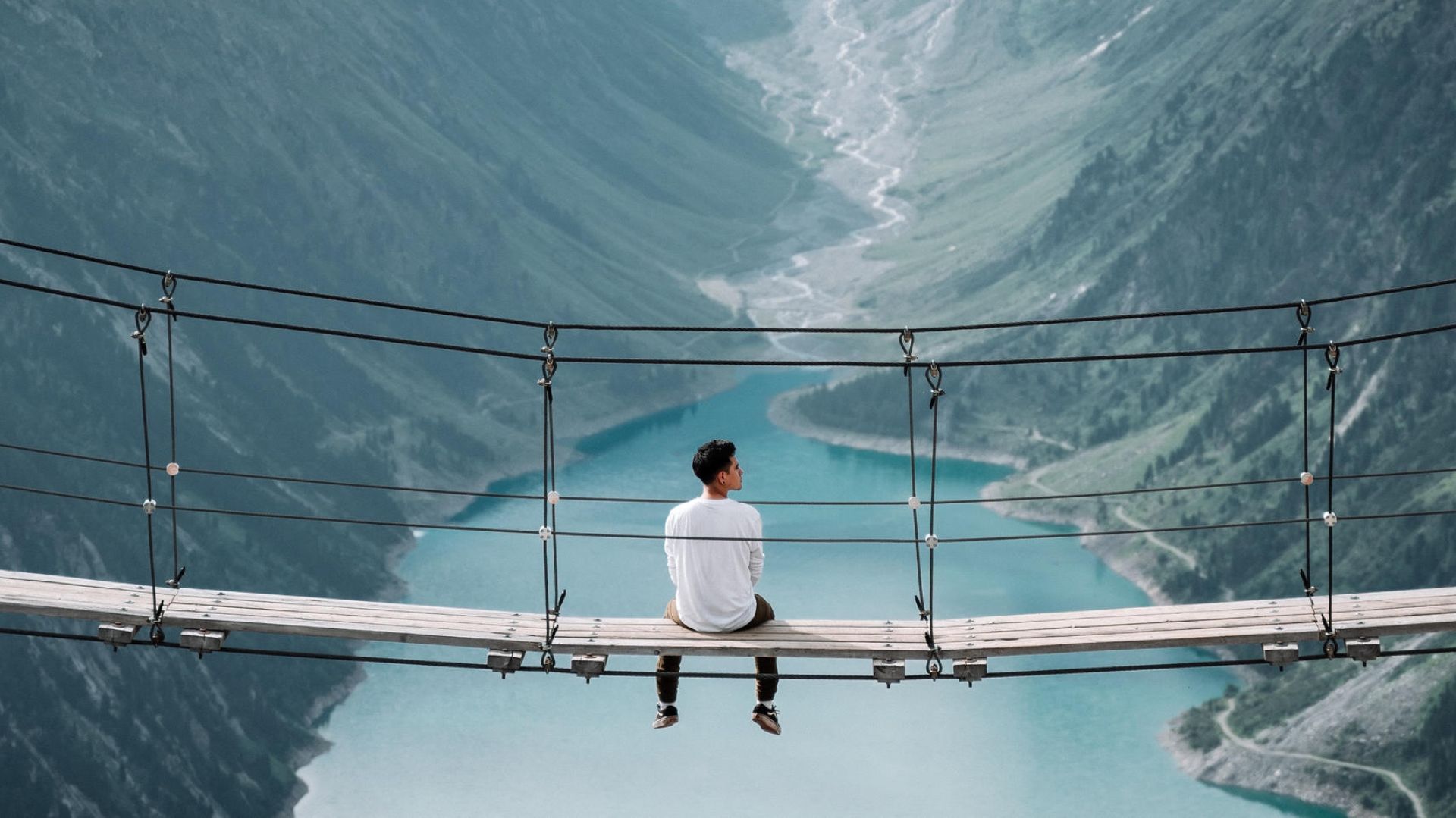 man sitting on a rope bridge looking out at the mountain