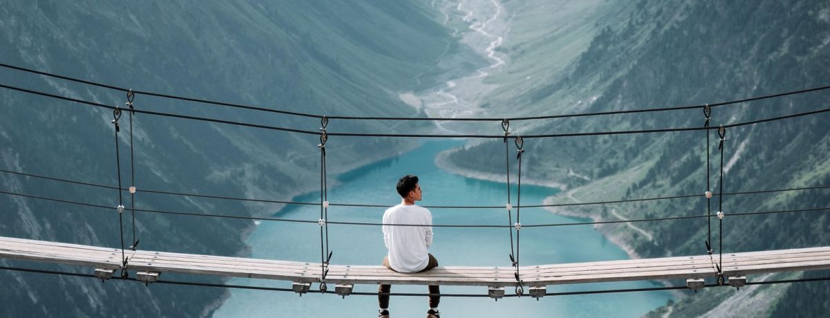 man sitting on a rope bridge looking out at the mountain