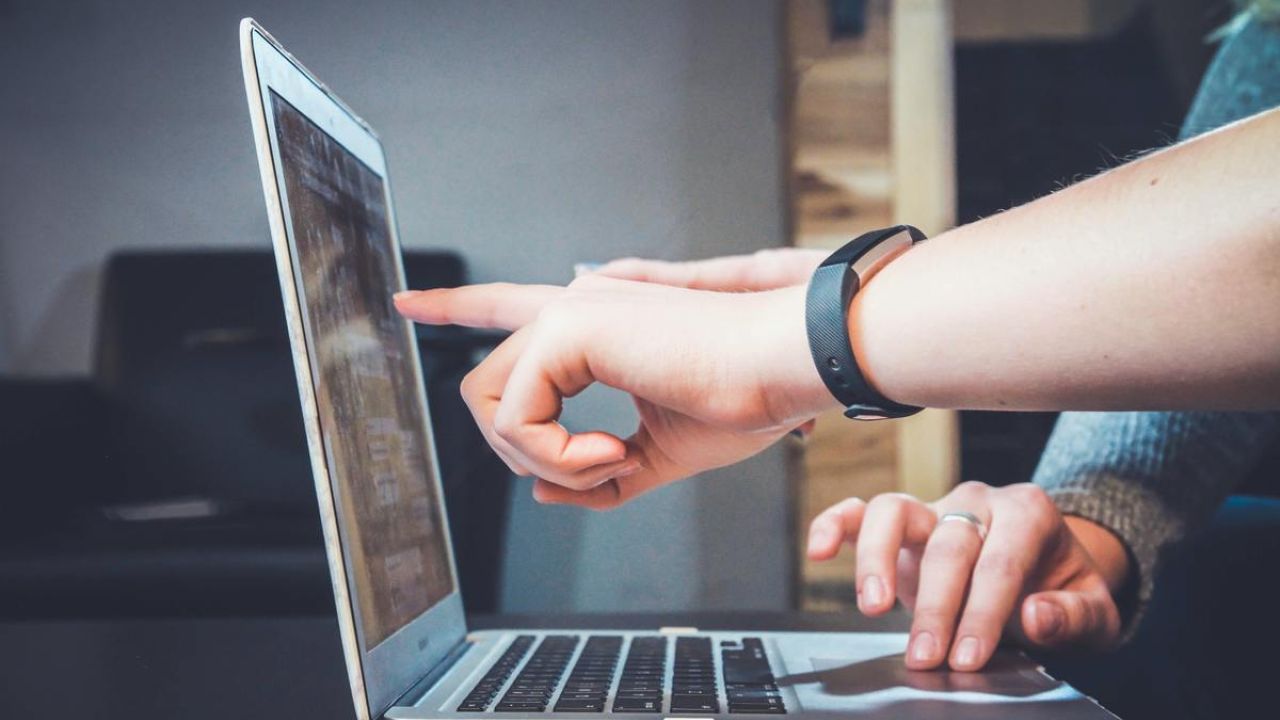 two people working together indoors at a desk pointing at a computer