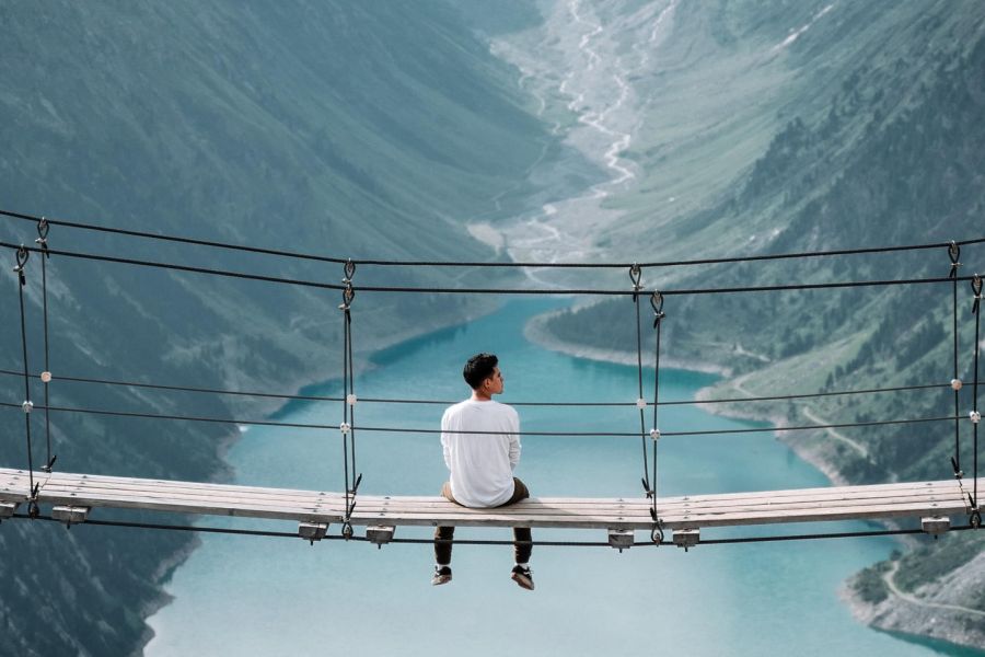 man sitting on a rope bridge looking out at the mountain