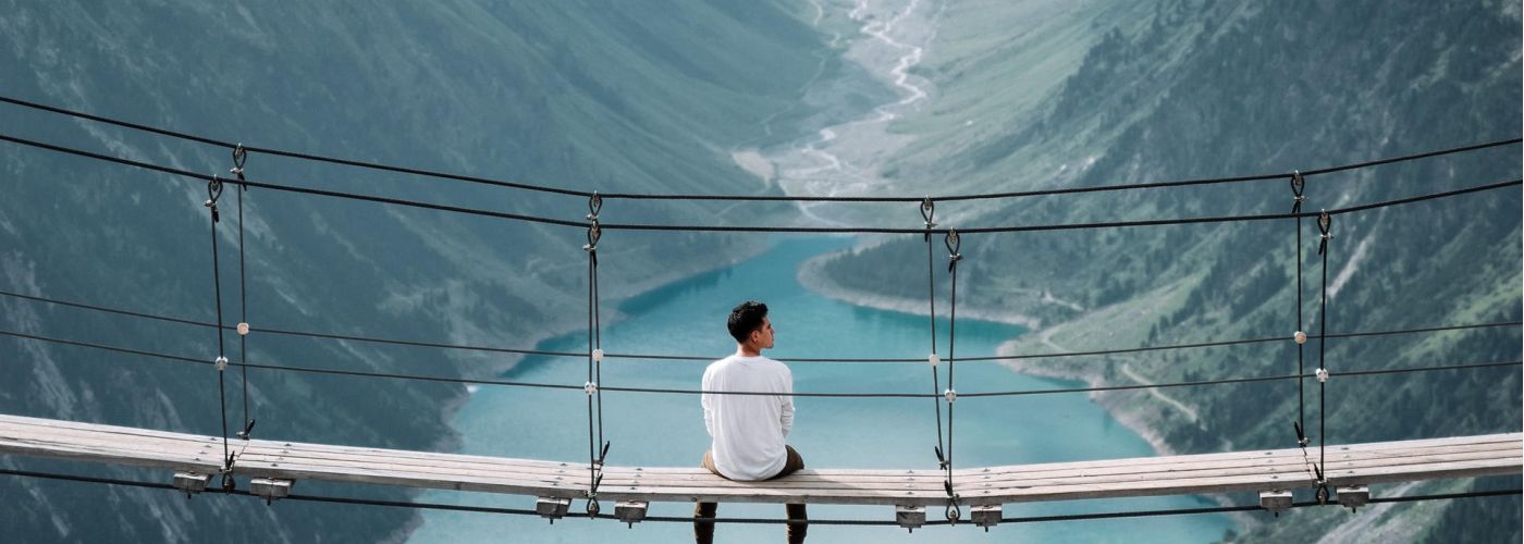 man sitting on a rope bridge looking out at the mountain